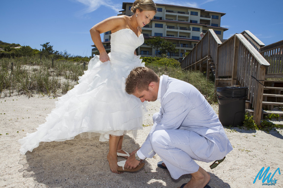 groom helping bride put her sandal on her foot on the beach