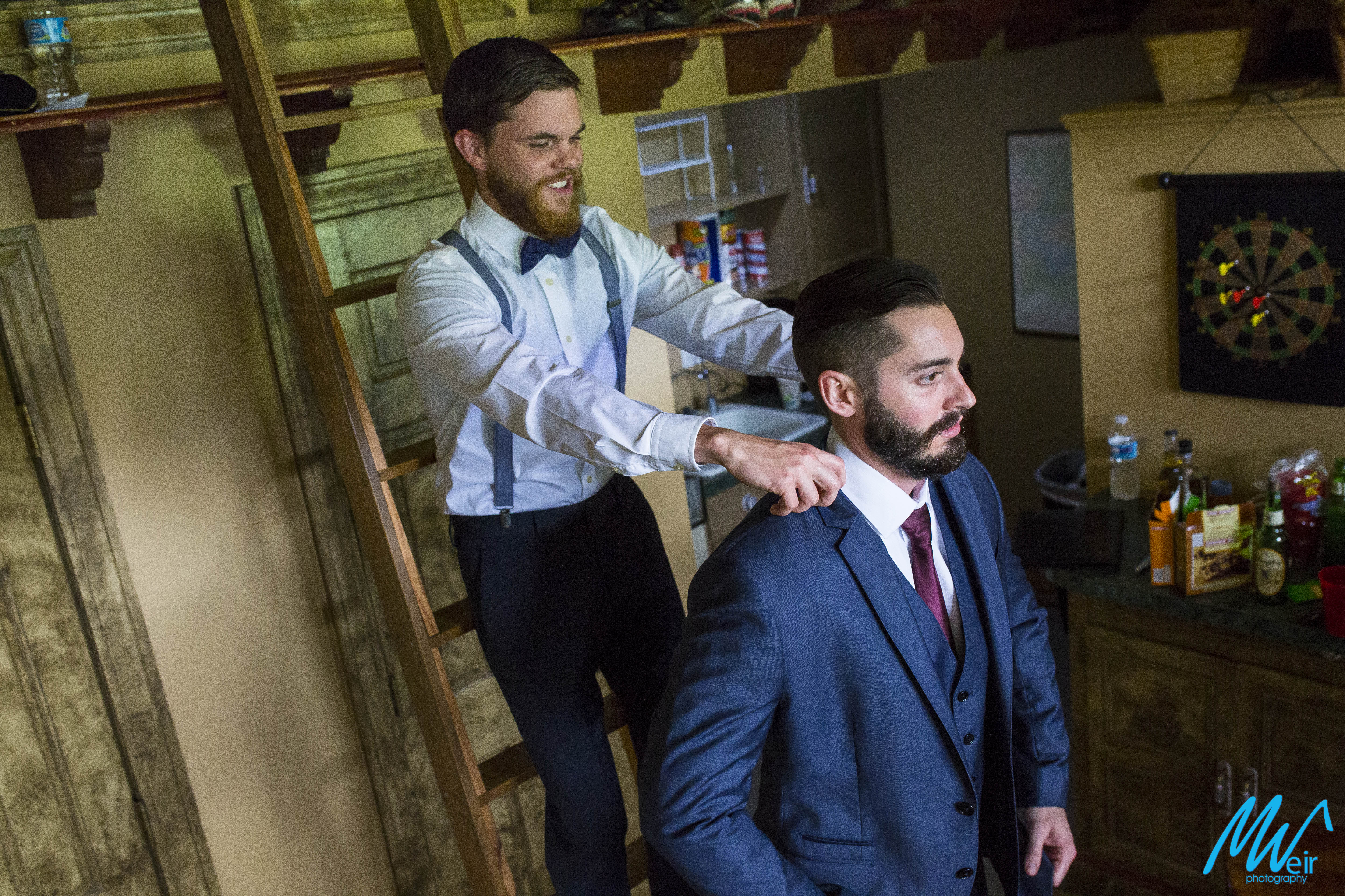 groomsman stands on a ladder to help groom put his jacket on