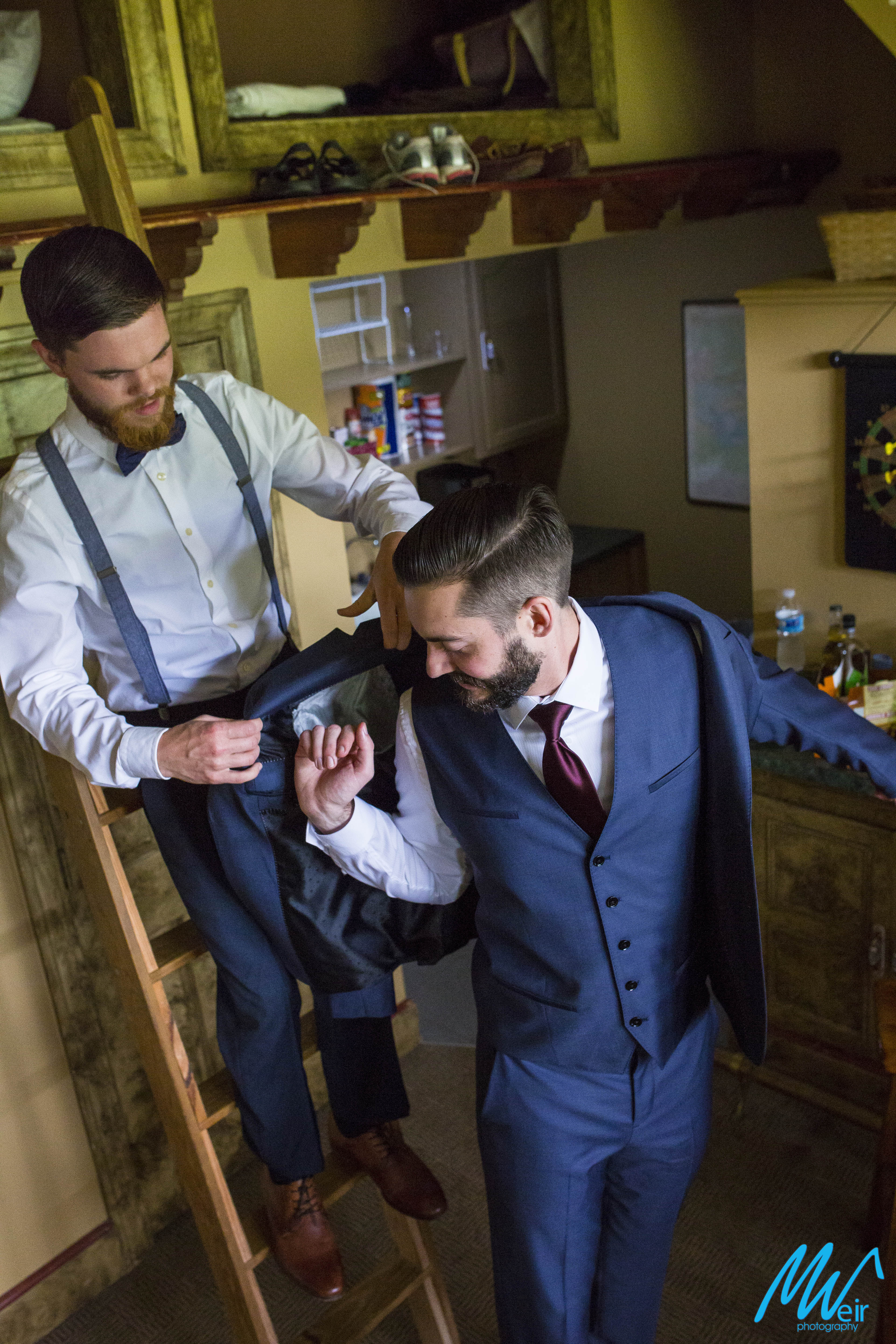 groomsman stands on a ladder to help groom put his jacket on