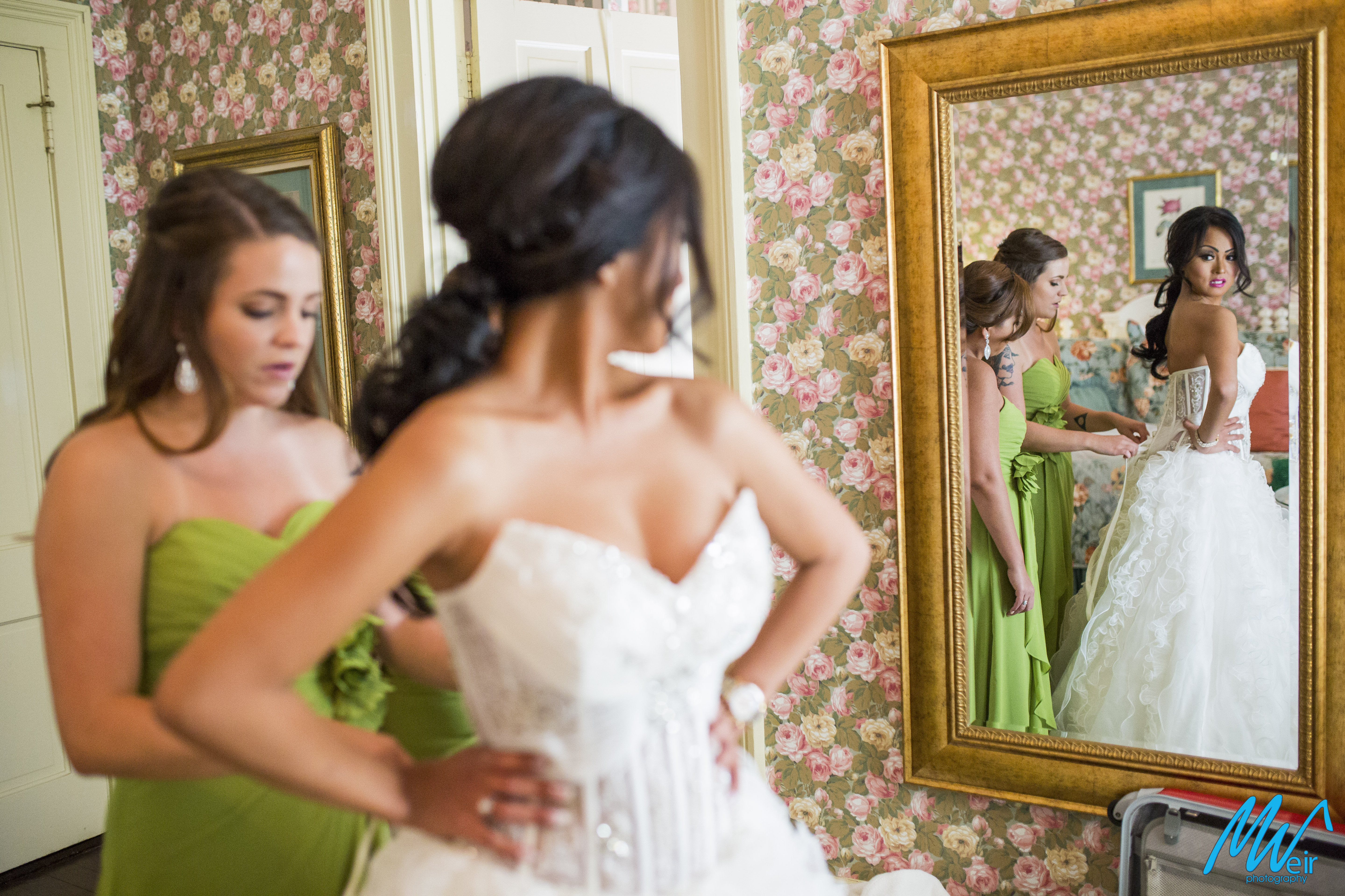 bride looks at herself in the mirror while the back of her dress is laced up