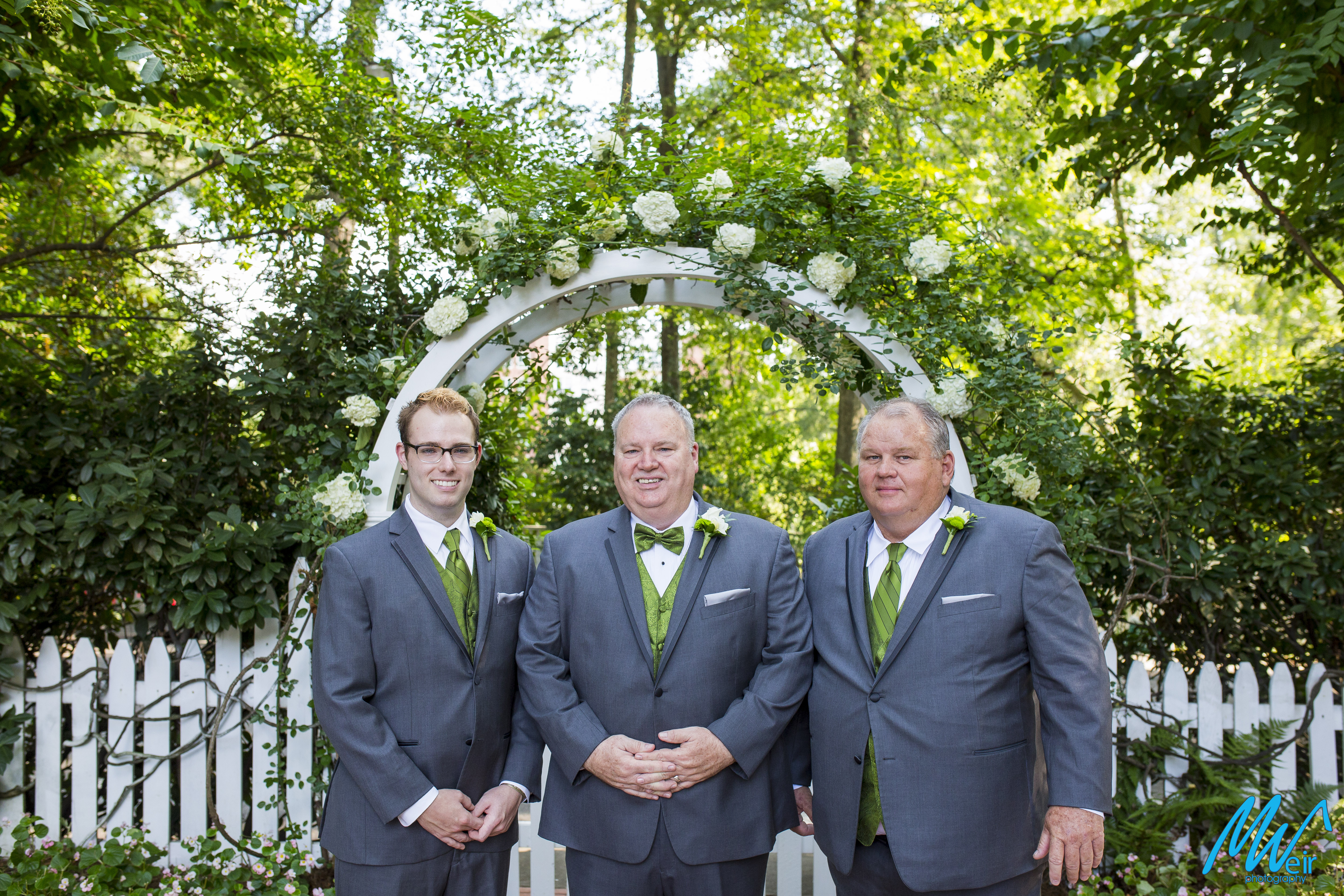 groom and groomsmen stand in front of garden arch