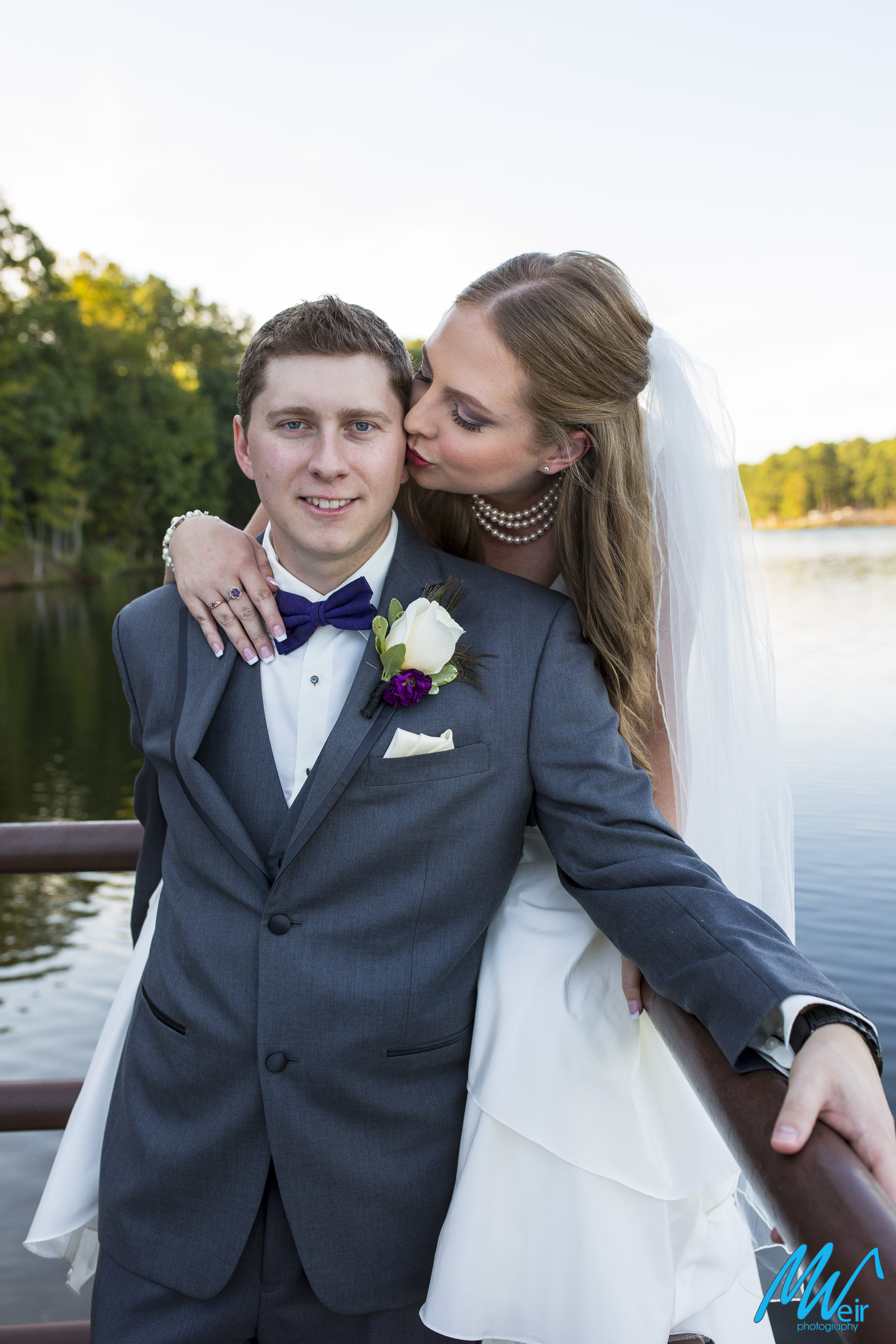 bride sits on railing next to a lake kissing grooms cheek