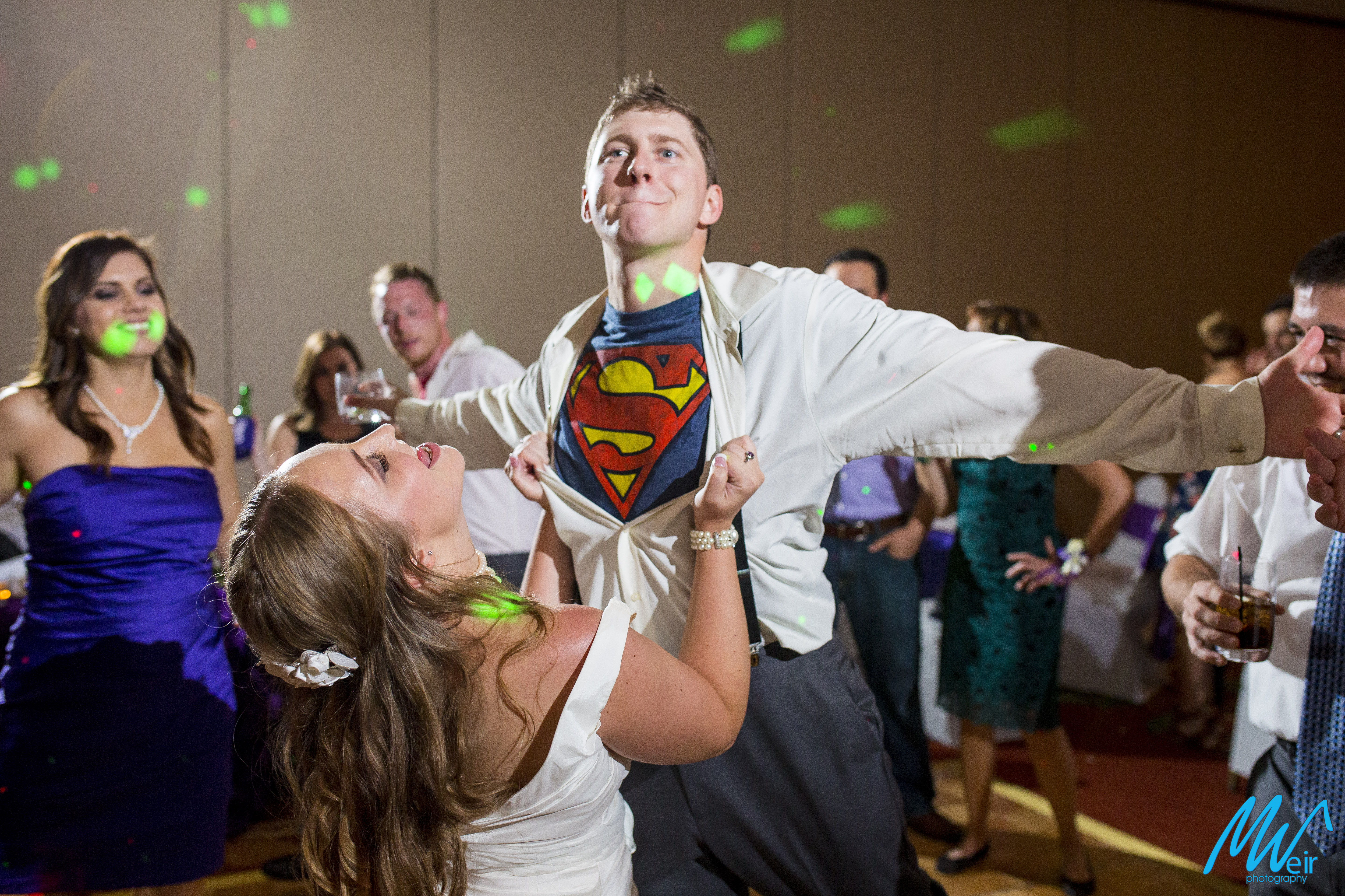 Bride opens grooms shirt to superman tshirt underneath while groom pretends to fly