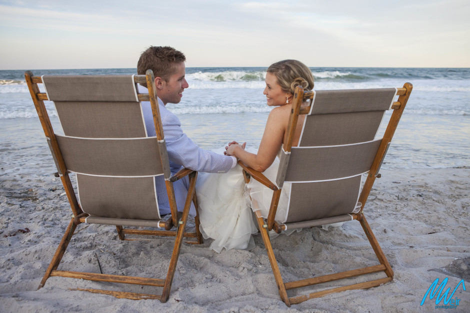 bride and groom holding hands on the beach while sitting in beach chairs