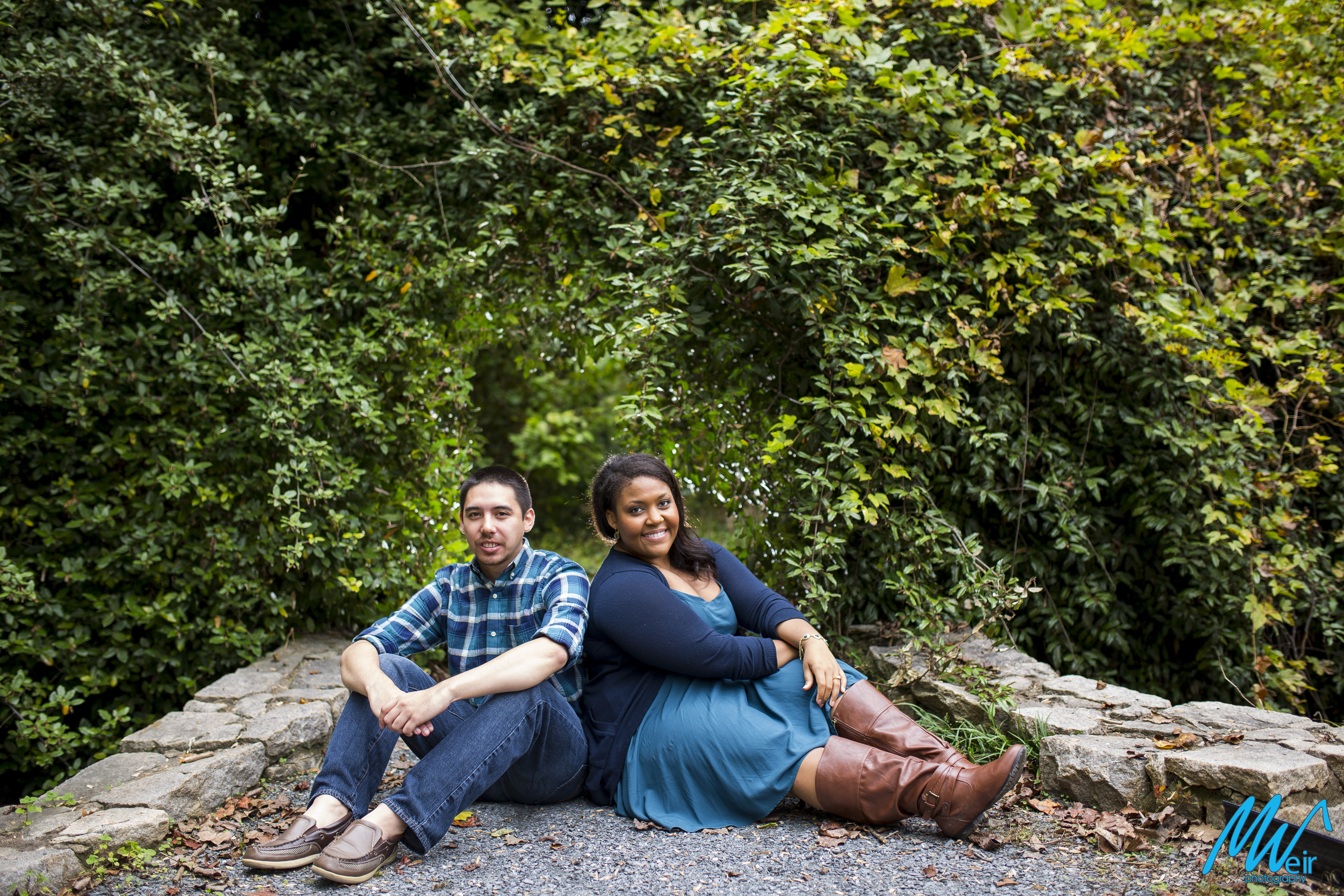 bride and groom sitting on a gravel path in front of over grown bush