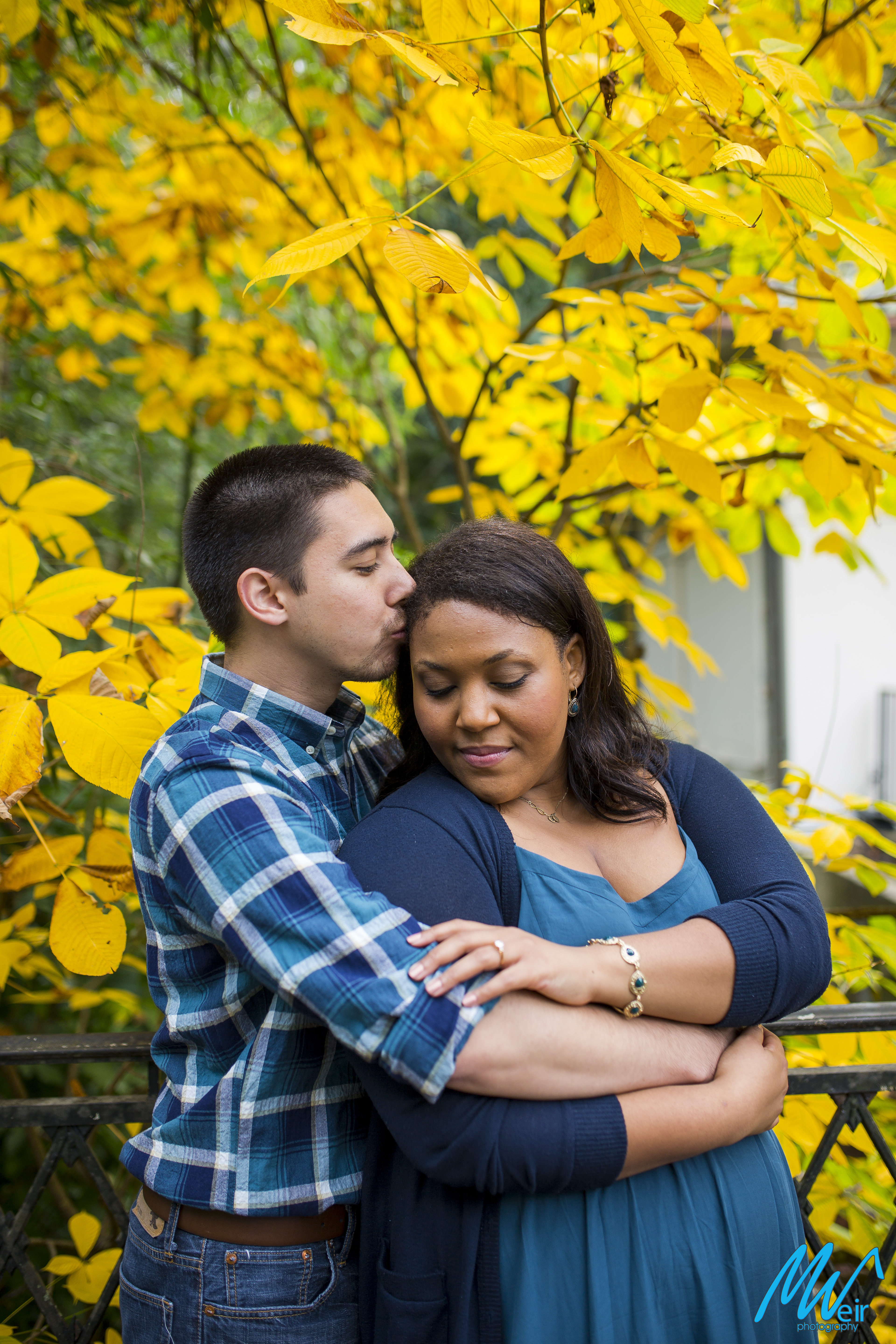 groom kisses bride's temple while holding her from behind in front of yellow fall leaves